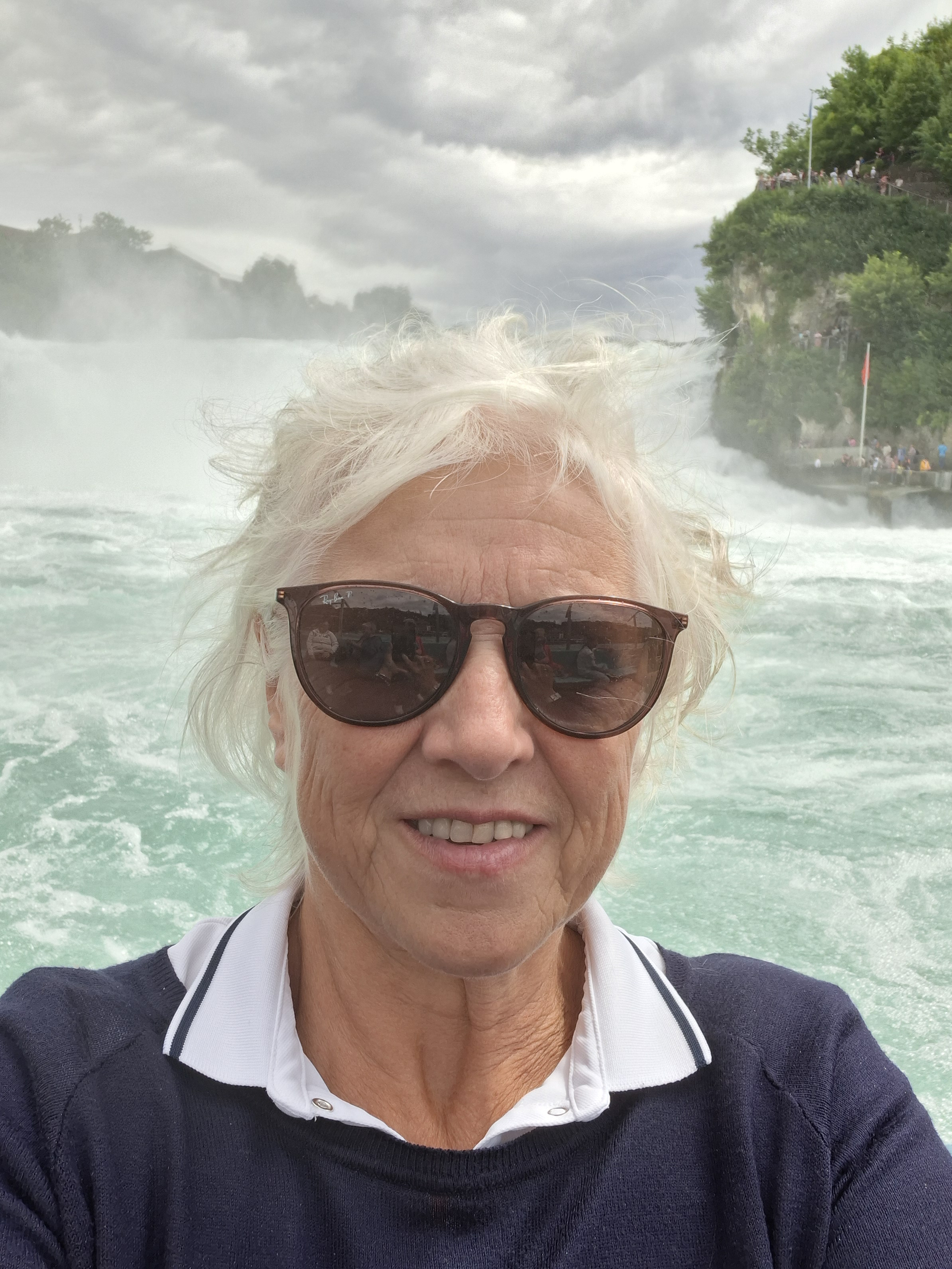 A woman with white hair and sunglasses stands in front of a body of water, possibly a river or lake, on a cloudy day. She ...